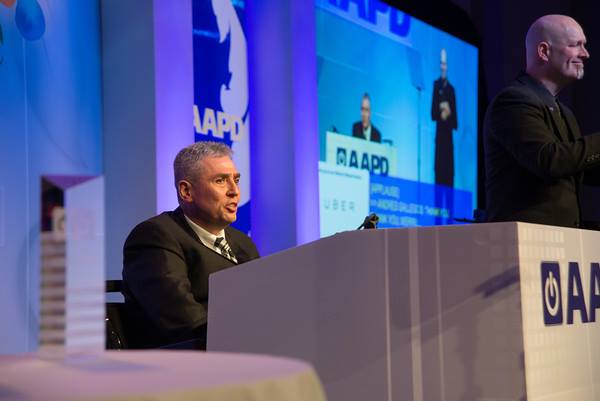 Andrés Gallegos, a Latino man with grey hair wears a suit and speaks to the audience from his wheelchair at the AAPD Leadership Awards Gala. He is standing on a stage with the AAPD logo and various purple and blue lighting. 
