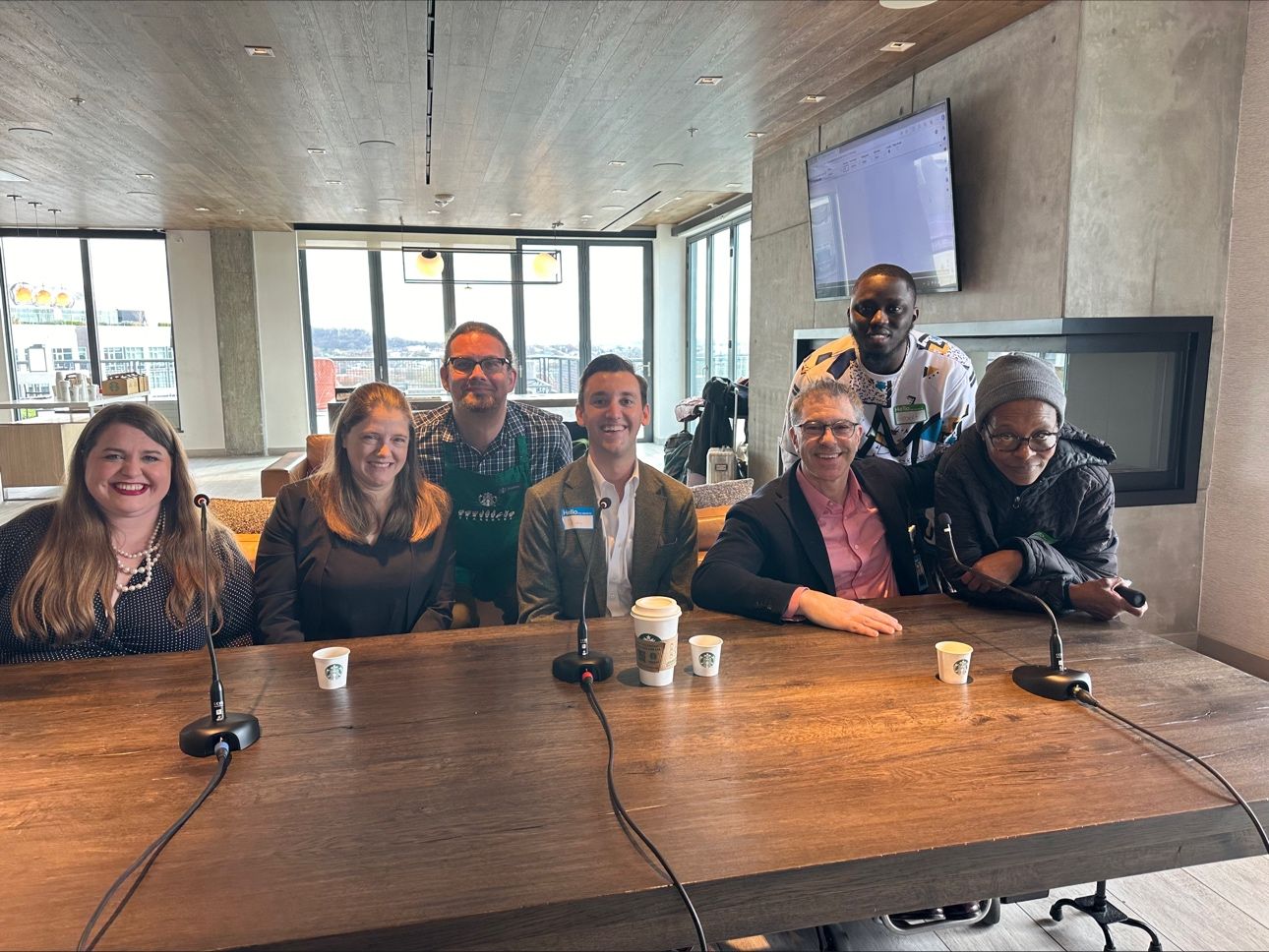 A group of seven people, six sitting and one standing behind them, posing for a photo at a long wooden table with microphones and Starbucks cups in front of them. The setting appears to be a modern, well-lit room with large windows and a screen on the wall in the background. Everyone is smiling and dressed in casual or business-casual attire.