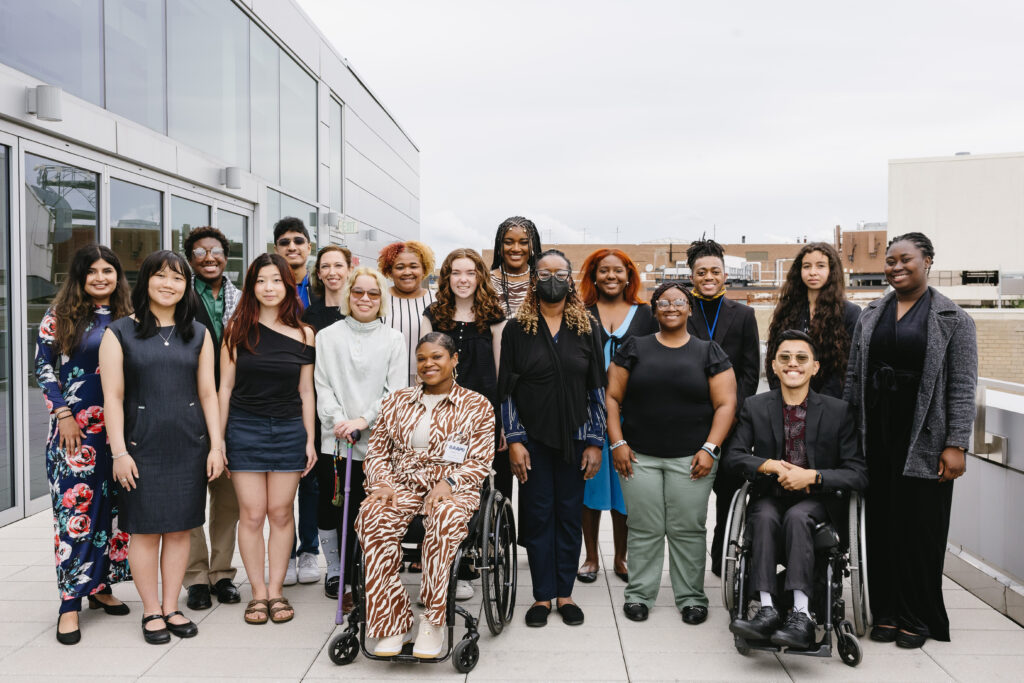 A group of AAPD interns smiles at the camera, gathered outside on a rooftop balcony. The group is a diversity of races and genders, and has a diversity of disabilities. Some members of the group are sitting in wheelchairs, others are standing, others are using canes. Some are wearing masks.