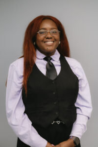Image Description: Alyssa, an African American woman in her 20s with long, straight reddish-brown hair and rectangular glasses, is smiling warmly at the camera and standing against a light gray background. They are wearing a light lavender dress shirt paired with a black vest and a black tie. They have long, straight reddish-brown hair and her hands are clasped in front of herself.