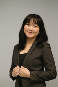 Charlotte, an East Asian woman in her 20s with long black hair and bangs, is smiling at the camera in front of a grey background. She is wearing a dark grey blazer and a black shirt with silver jewelry. 