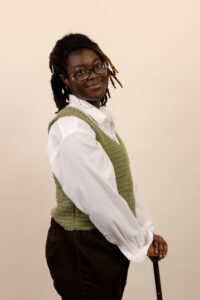 Ahna is a Black person with mostly black and bleached locs. He is wearing a white shirt with a green vest. He is standing to the side holding his cane and smiling.