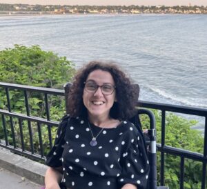 A woman with curly brown hair and glasses smiles at the camera while seated in her wheelchair.