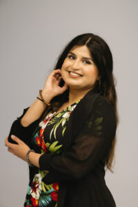 Tooba, a South Asian woman with light tan skin and long, wavy brown hair, smiles at the camera. She was one arm crossed across her body and the other resting under her chinm and is wearing a floral top and a black blazer.  