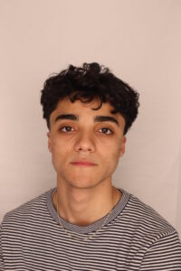 ID: Kemper has curly black hair and is wearing a striped shirt and a gold chain necklace against a plain, light-colored background.