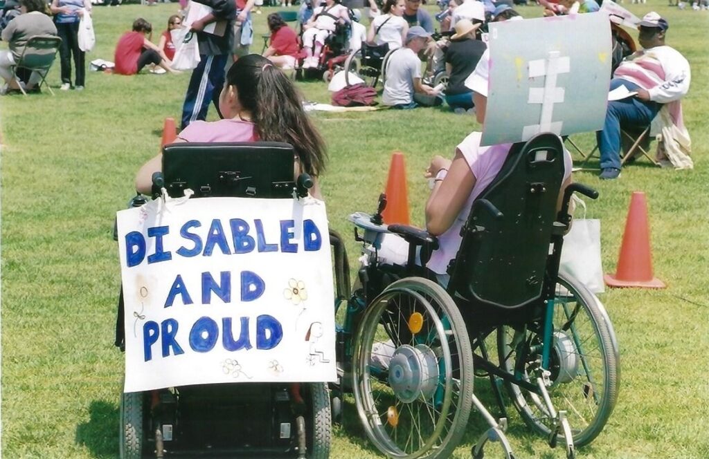Two wheelchair users at a protest with their backs turned to the camera. One of them has a sign visible on the back of their chair.