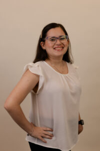 A young woman standing in front of a peach-colored wall. She has straight, dark brown hair and is wearing glasses. She is smiling and has light skin. She is wearing a white top. She is also wearing a silver necklace.