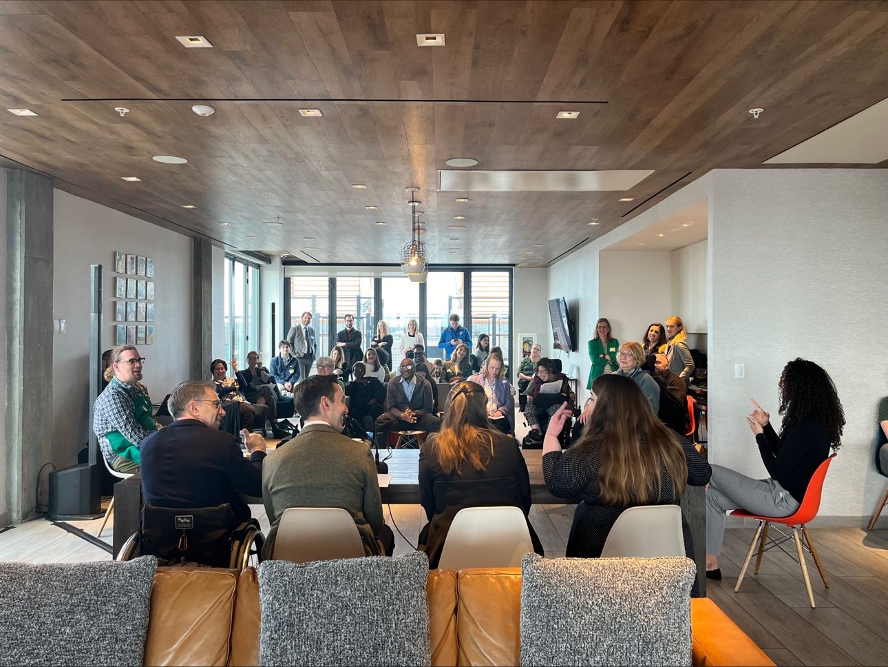 A group of people gathered in a modern room for a discussion or presentation. The photo is taken from behind four panelists sitting at a long table with microphones, facing an audience seated and standing in the background. The room is well-lit with large windows, a wooden ceiling, and a screen on the wall. The atmosphere appears engaged and attentive, with diverse individuals participating.
